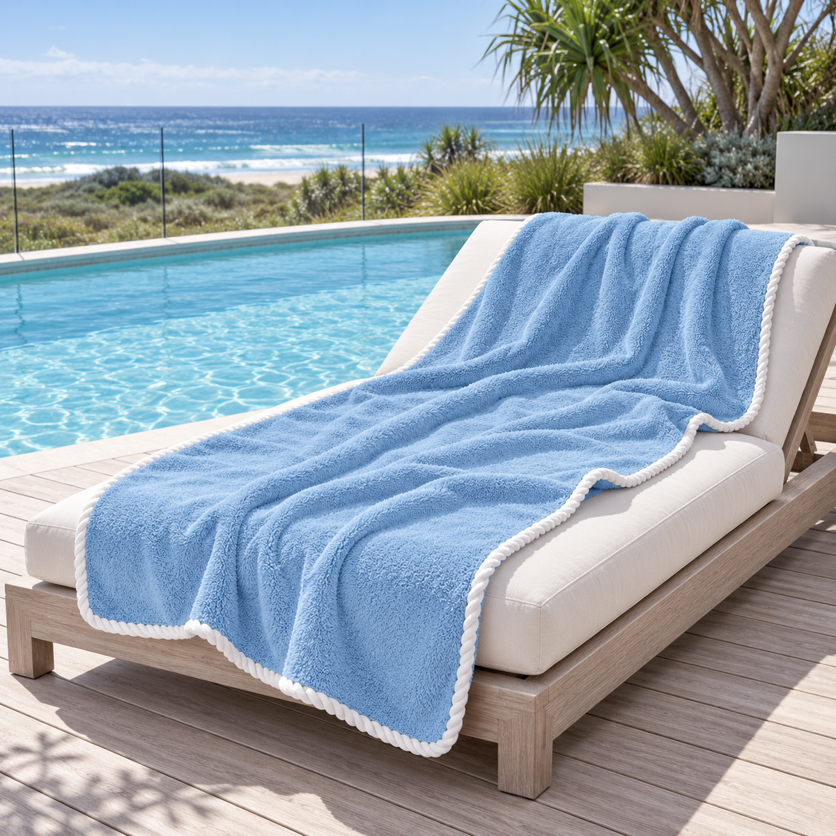 Noosa extra large light blue towel with white piped edging draped over a timber poolside lounger overlooking an Australian coastal beach bay.