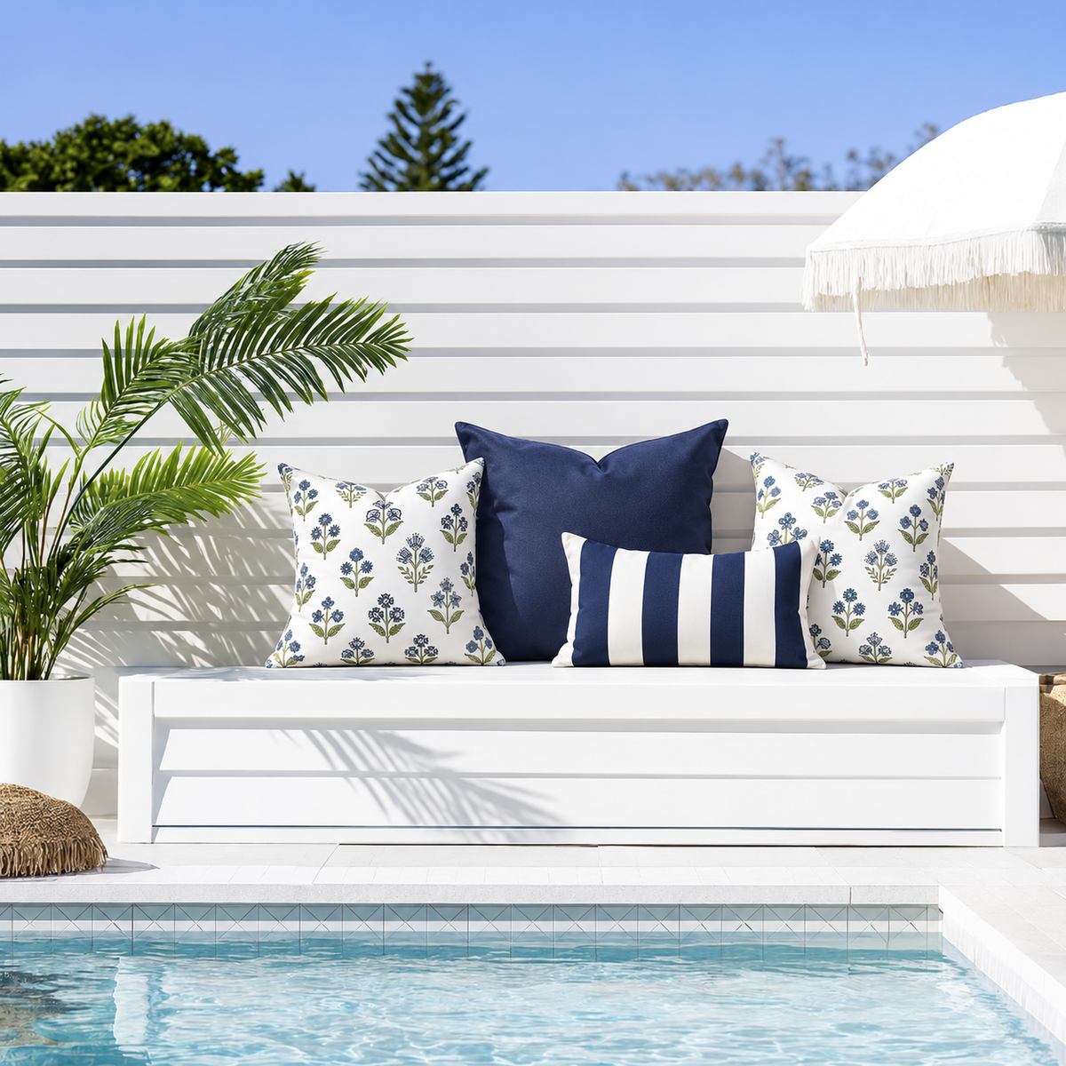 Four weather-resistant outdoor cushion covers Hamptons styled on a white poolside bench: two Fleur blue and green floral 45 x 45 cm cushions, one large navy 60 x 60 cm cushion and one navy and white striped 30 x 50 cm rectangular cushion.