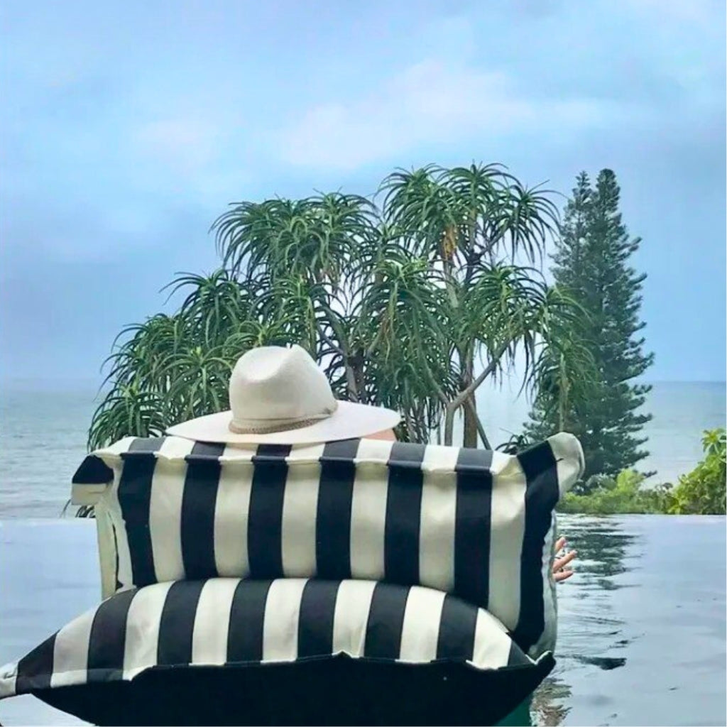Lifestyle image of the Big Bertha single floating beanbag lounger cover in black and white stripe, positioned in an infinity pool overlooking the water.