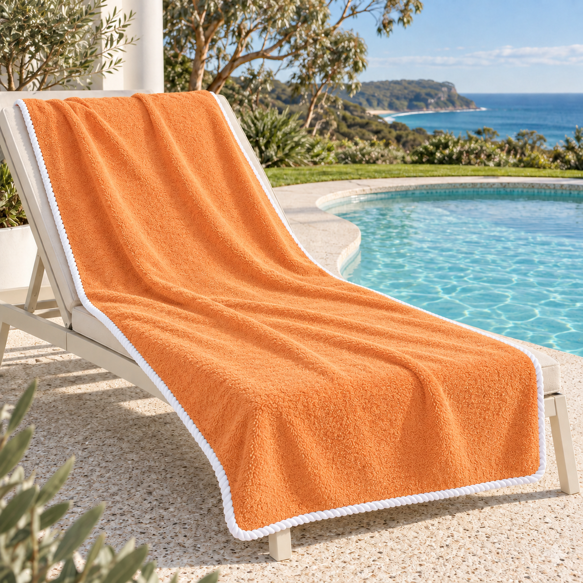 Noosa extra large tangerine towel with white piped edging on a poolside lounger overlooking an Australian coastal headland.